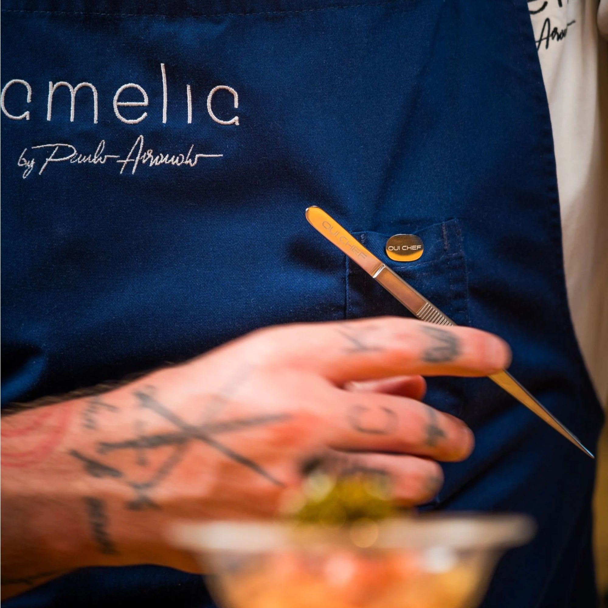 Chef wearing a blue apron with 'Amelia' branding attaching small stainless steel tweezers to the holdfast magnetic clip