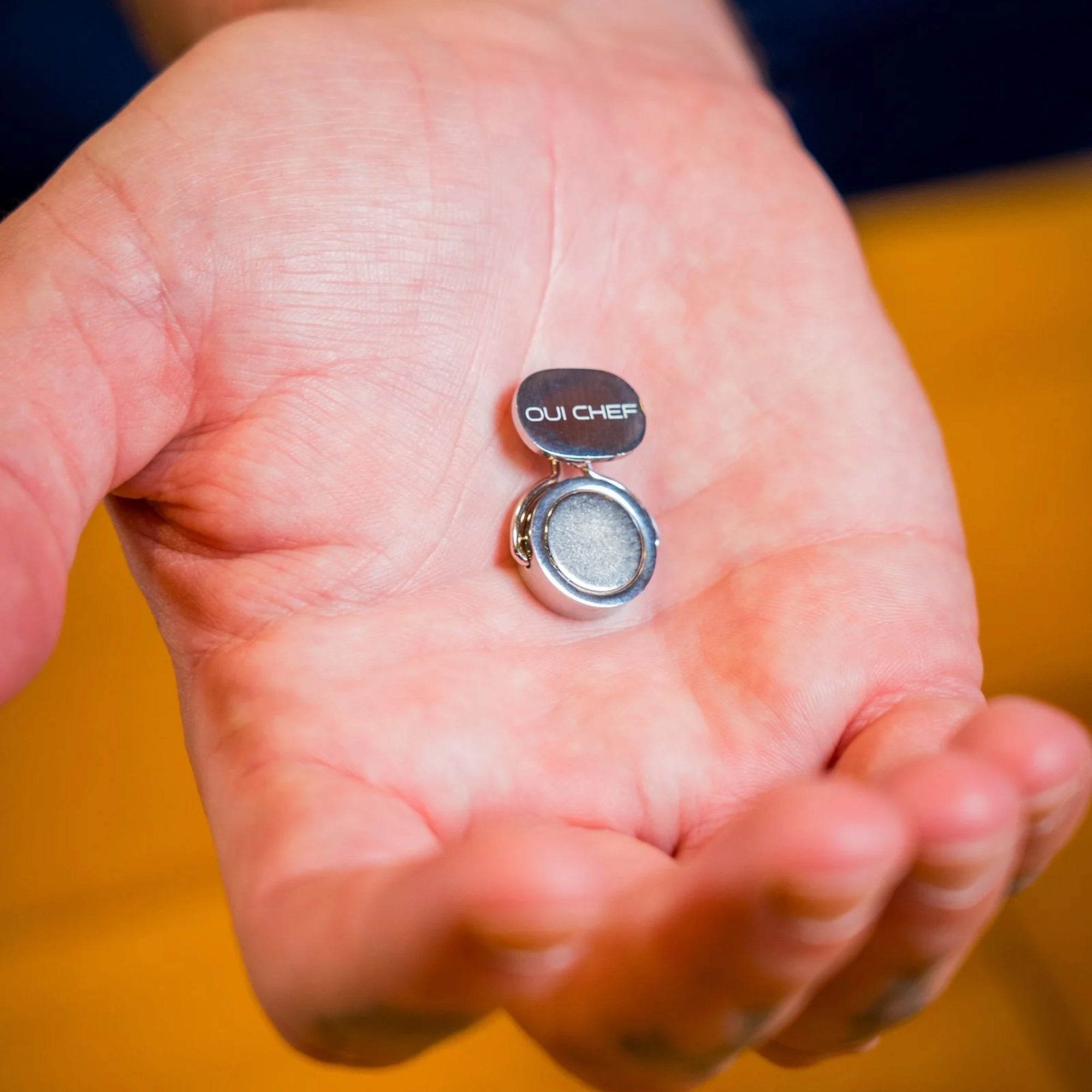 Holdfast magnetic clip with 'Oui Chef' branding held in a chef's hand against a blurred background