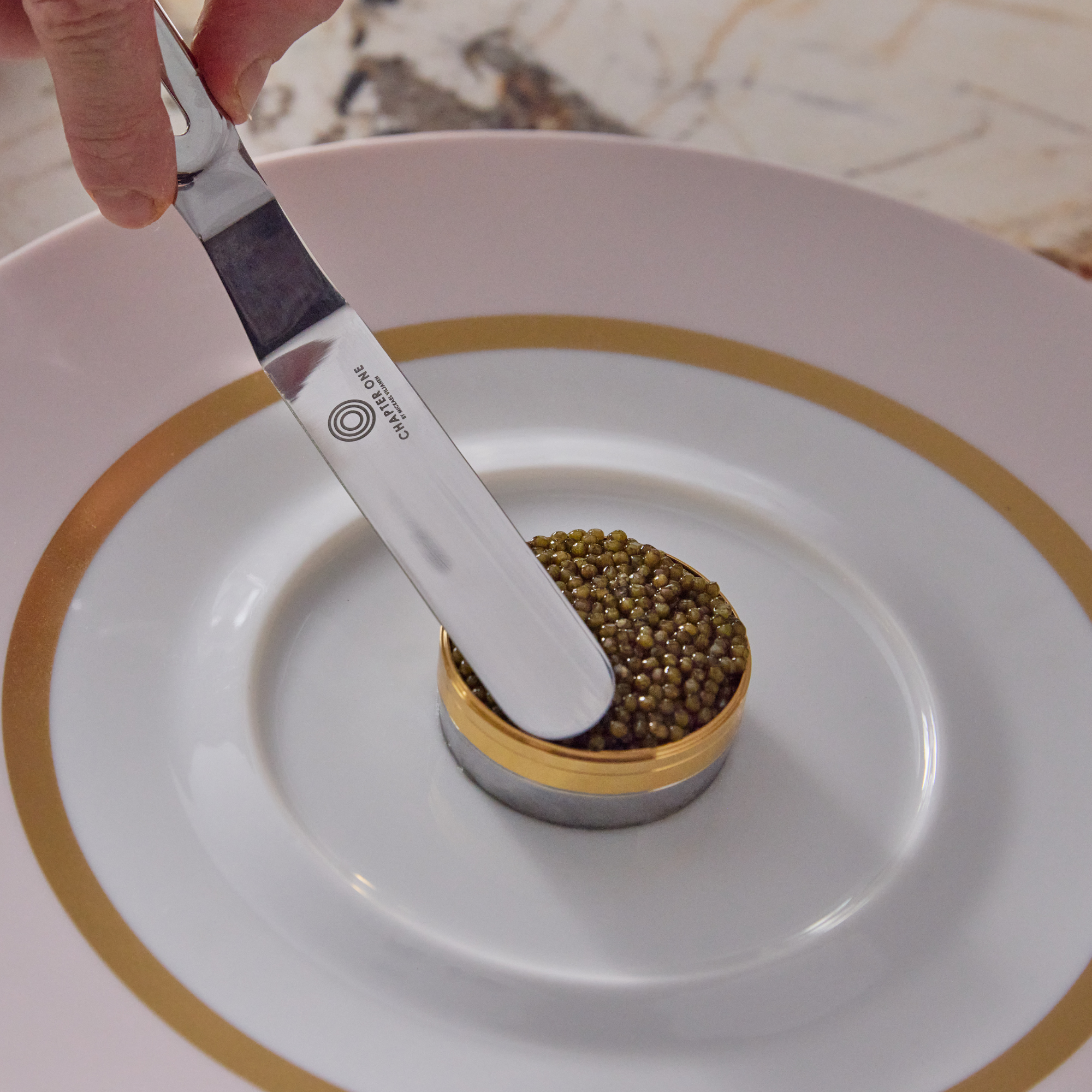 Chef delicately smoothing over caviar that is sitting in a ring mould on a white plate with a stainless steel palette knife that has "Chapter One" logo engraved on it.