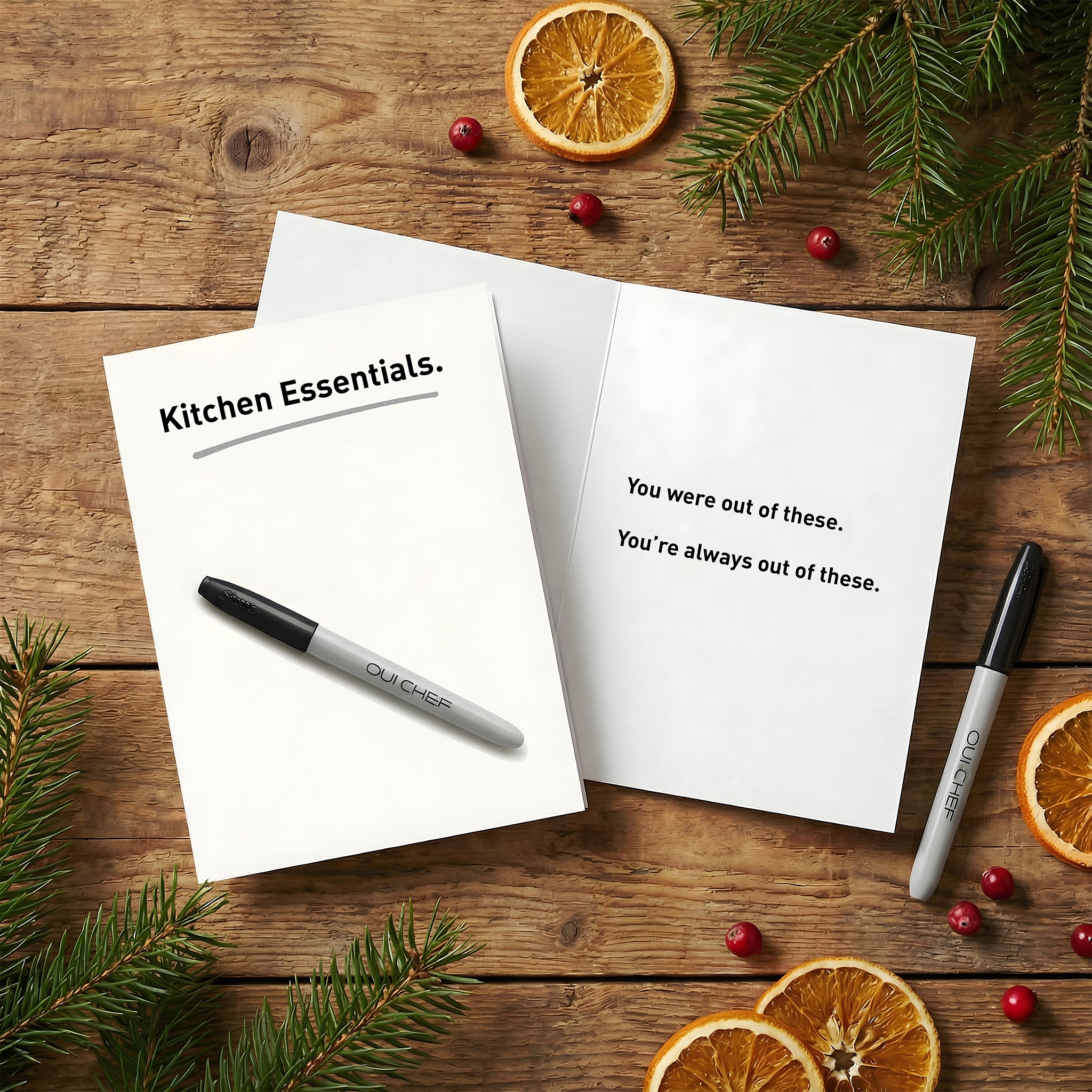 Oui Chef "Kitchen Essentials" Greeting card laid flat on a kitchen table surrounded by festive greenery - showing the front and inside of the card.