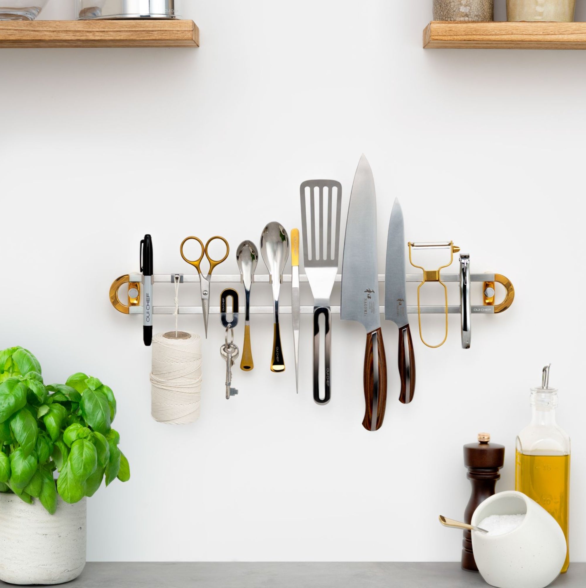 A Stainless steel magnetic rack placed on a white home kitchen wall with the following line up from left to right; a sharpie, twine, scissors, a set of keys, two spoons, chef's tweezers, a stainless spatula, 2 kitchen knives, a gold peeler and a chrome corkscrew.