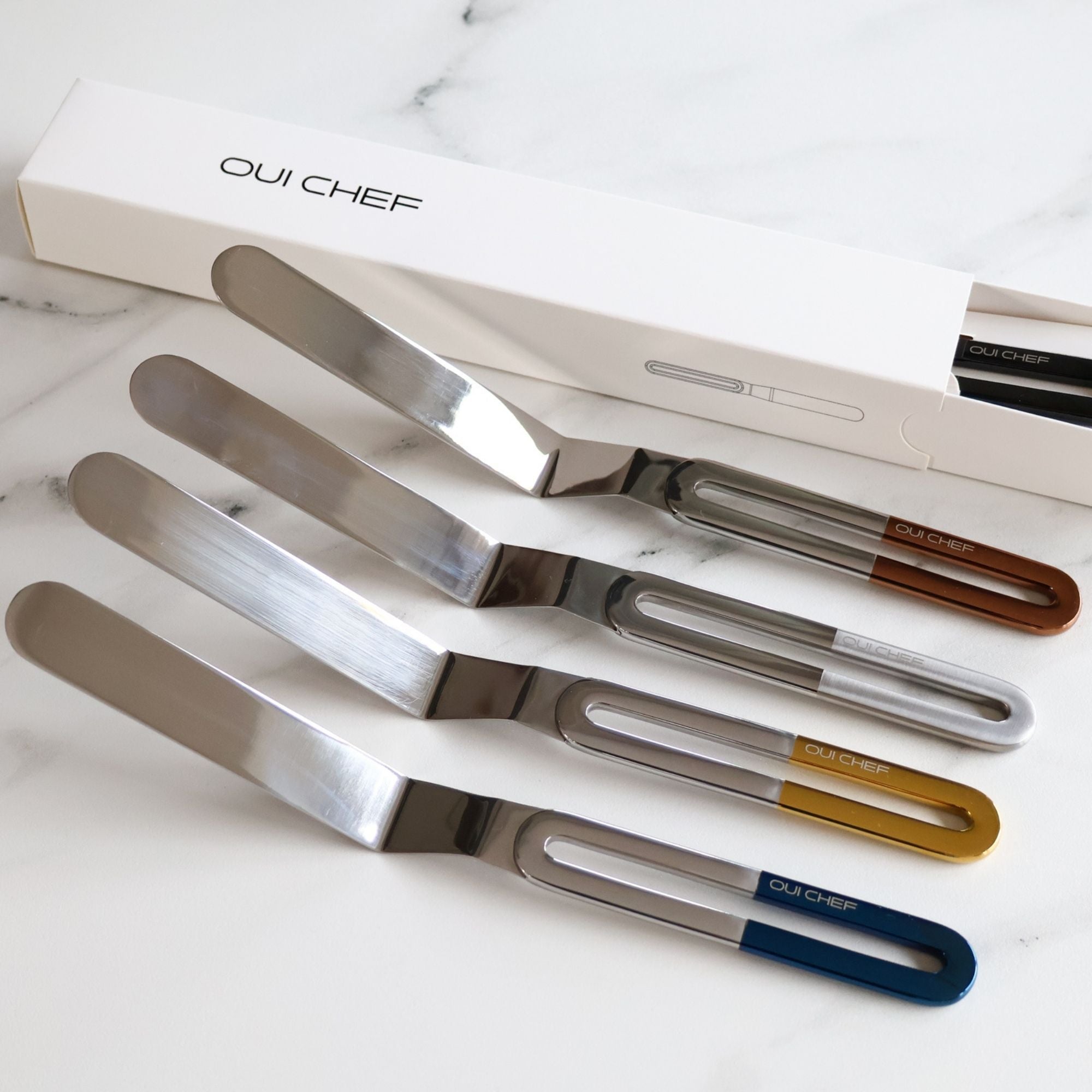 Four stainless steel offset palette knives of the larger size, each with a sleek metal blade and handles in chrome, gold, metallic blue and copper with an ergonomic handle engraved “OUICHEF,” arranged side by side on a marble worktop. 