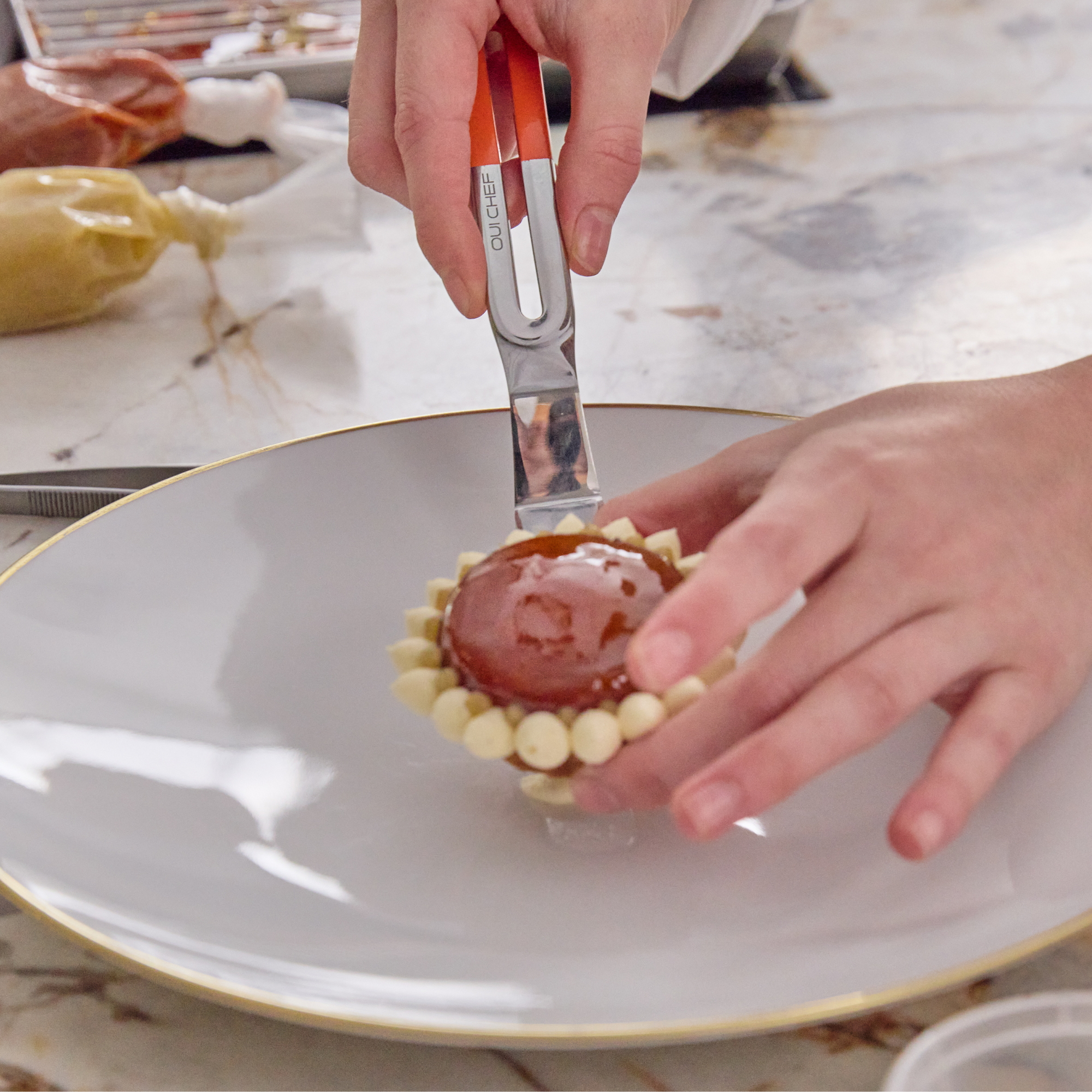 Chef lifting and delicately placing a dessert tart on to a white plate with a stainless steel palette knife that has an edge-to-edge sharp blade and powdered orange colour handle.