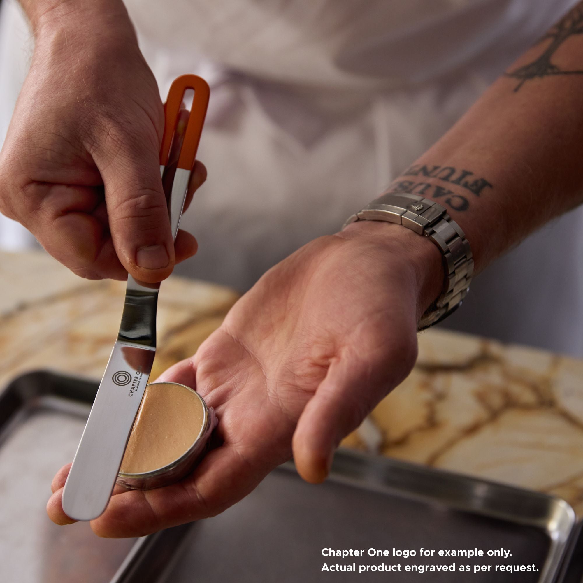 Chef smoothing a tin of pate with a stainless steel palette knife that has an edge-to-edge sharp blade and powdered orange colour handle with the "Chapter One" logo engraved on it.